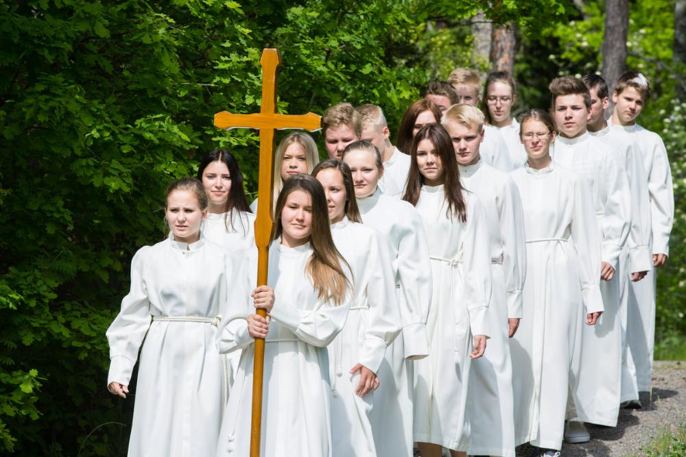 Joukko valkoiseen kaapuun pukeutuneita nuoria parijonossa. Ensimmäinen kanssa puista ristiä.