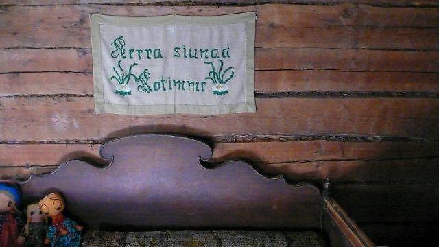 Vanha puinen sohva, yläpuolella kangas, jossa teksti: Herran siunaa kotimme