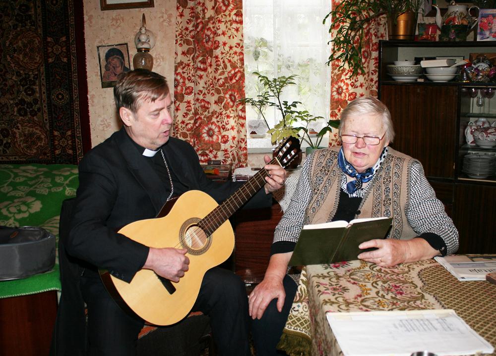 Miespappi soittaa kitaraa ja laulaa virsikirjasta vanhan naisen kanssa.