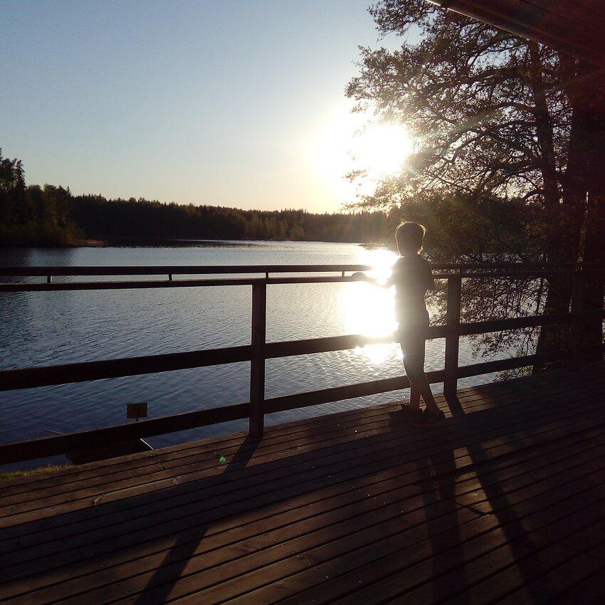 Auringonlasku järvellä. Etualalla poika nojaa laiturin kaiteeseen