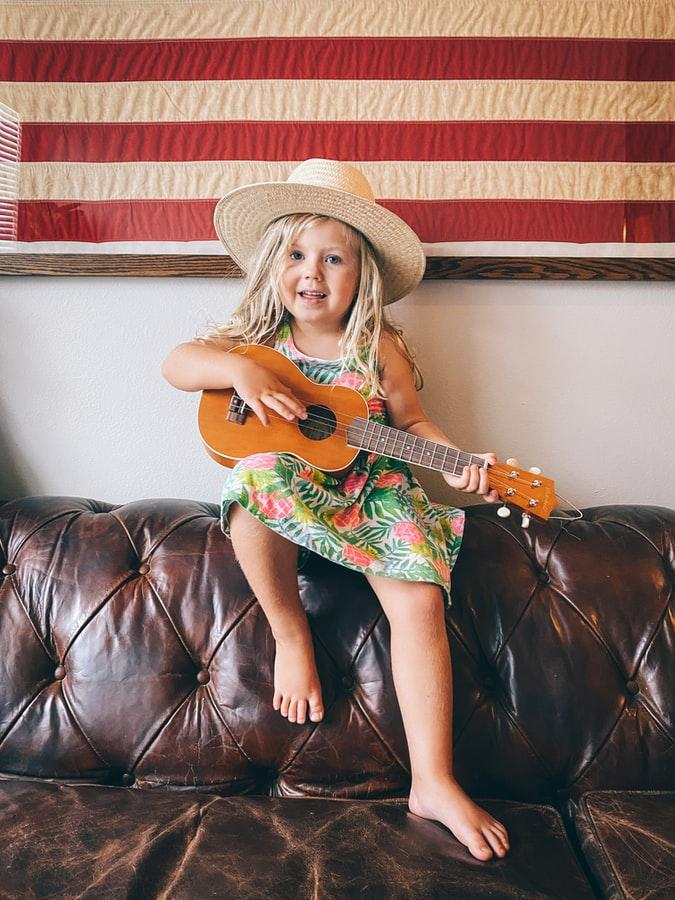 Hattupäinen tyttö soittaa ukulelea sohvan selkänojalla istuen