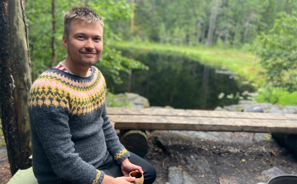 Matti Hernesaho istuu kuksa kädessä nuotiopaikalla lammen lähellä.